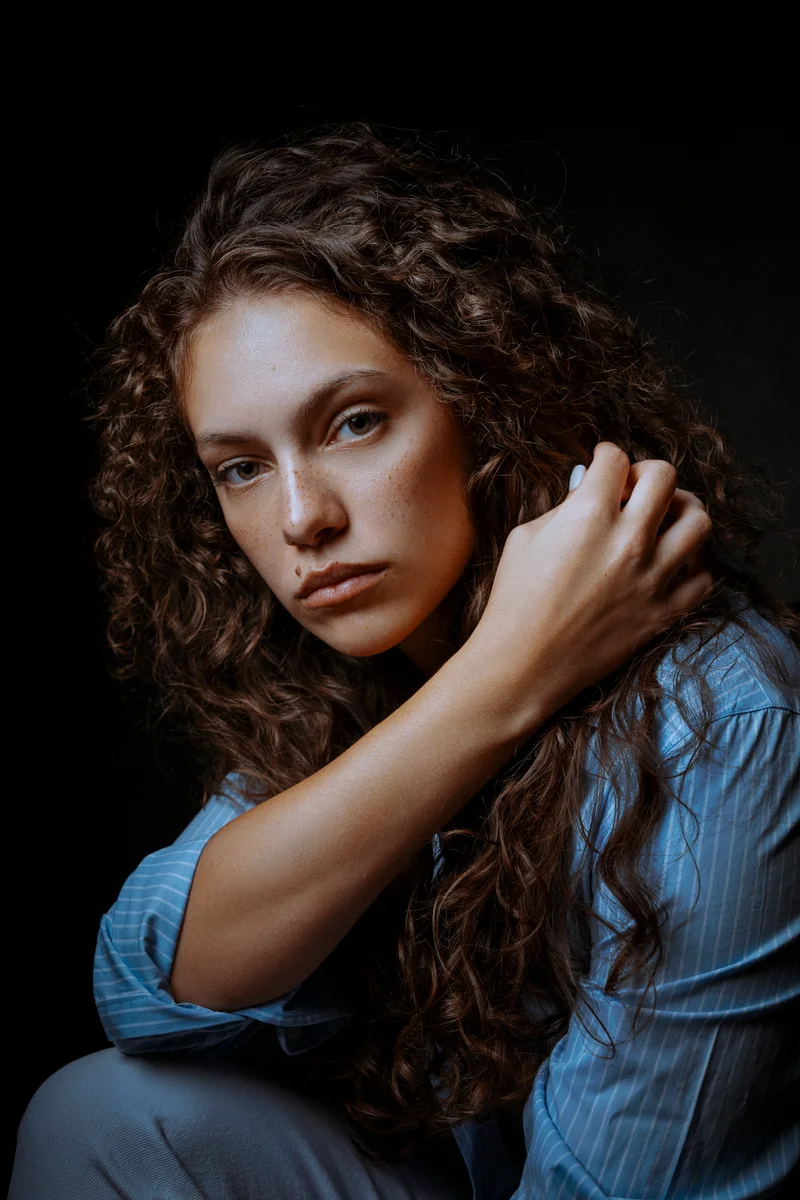 Woman with long curly hair in a blue striped shirt, her hand on her shoulder