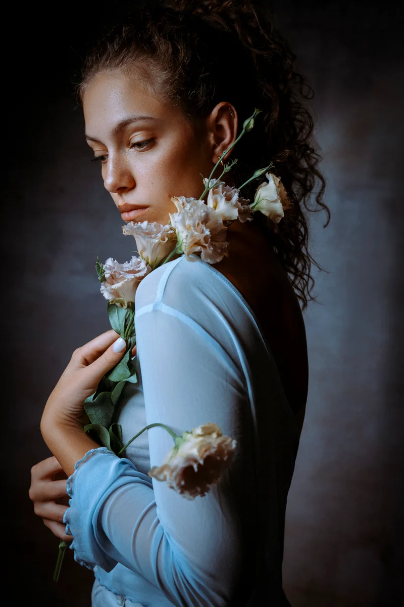 Woman with curly hair pinned up in a pale blue top, holding white flowers, looking down