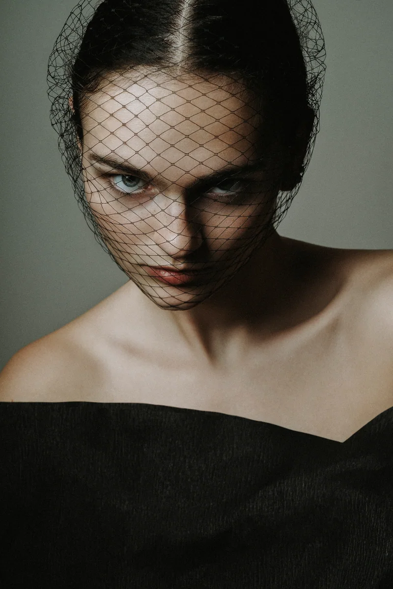 Woman behind a black net veil looking up from under her brows in soft studio light