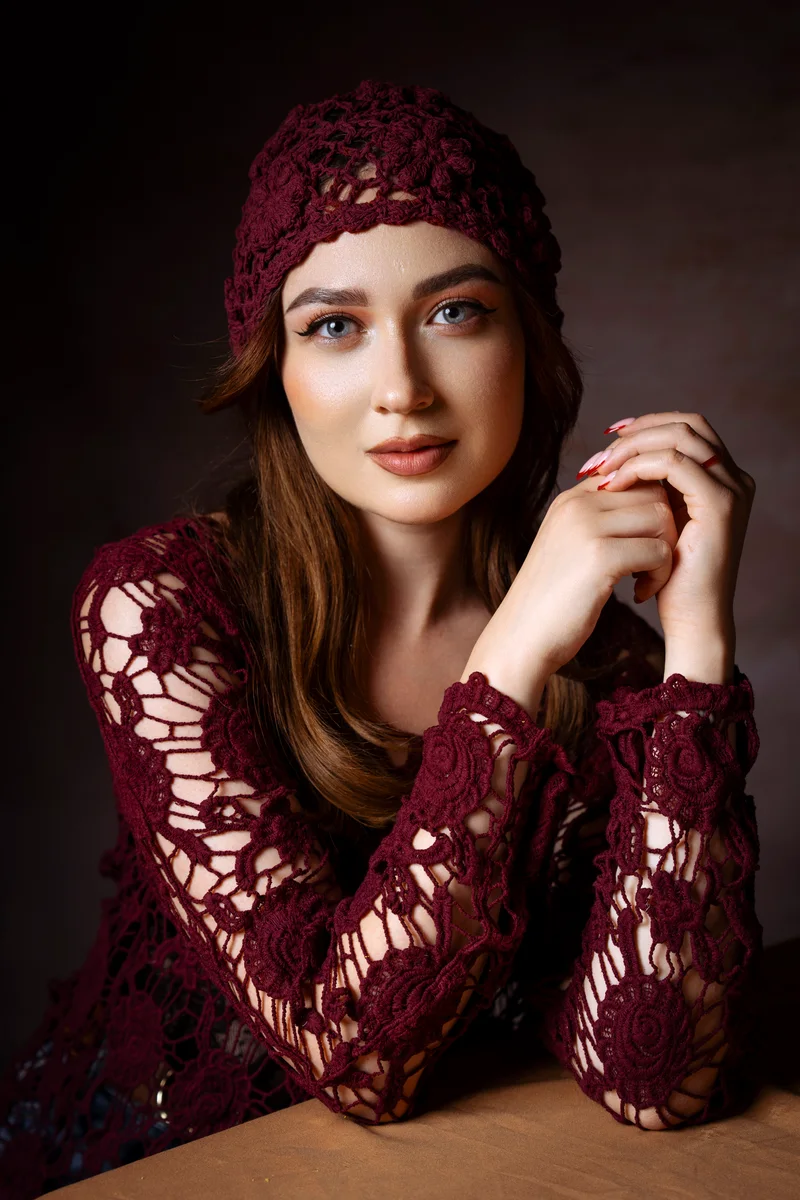Woman in a burgundy crochet cap leaning on a table with clasped hands