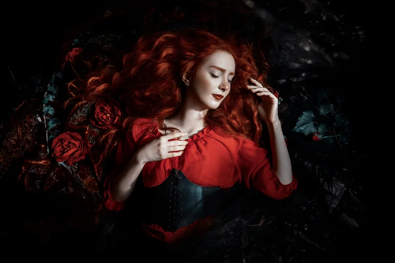 Red-haired woman in a red dress and black corset lying among dark roses
