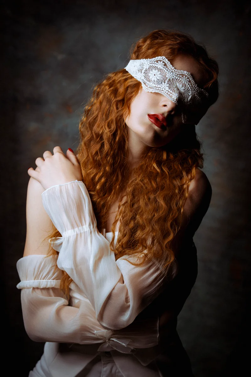 Woman with long curly red hair, a lace blindfold and a white blouse