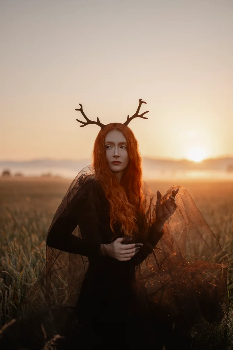 Red-haired woman with antlers and a black tulle dress in a field against a low sun