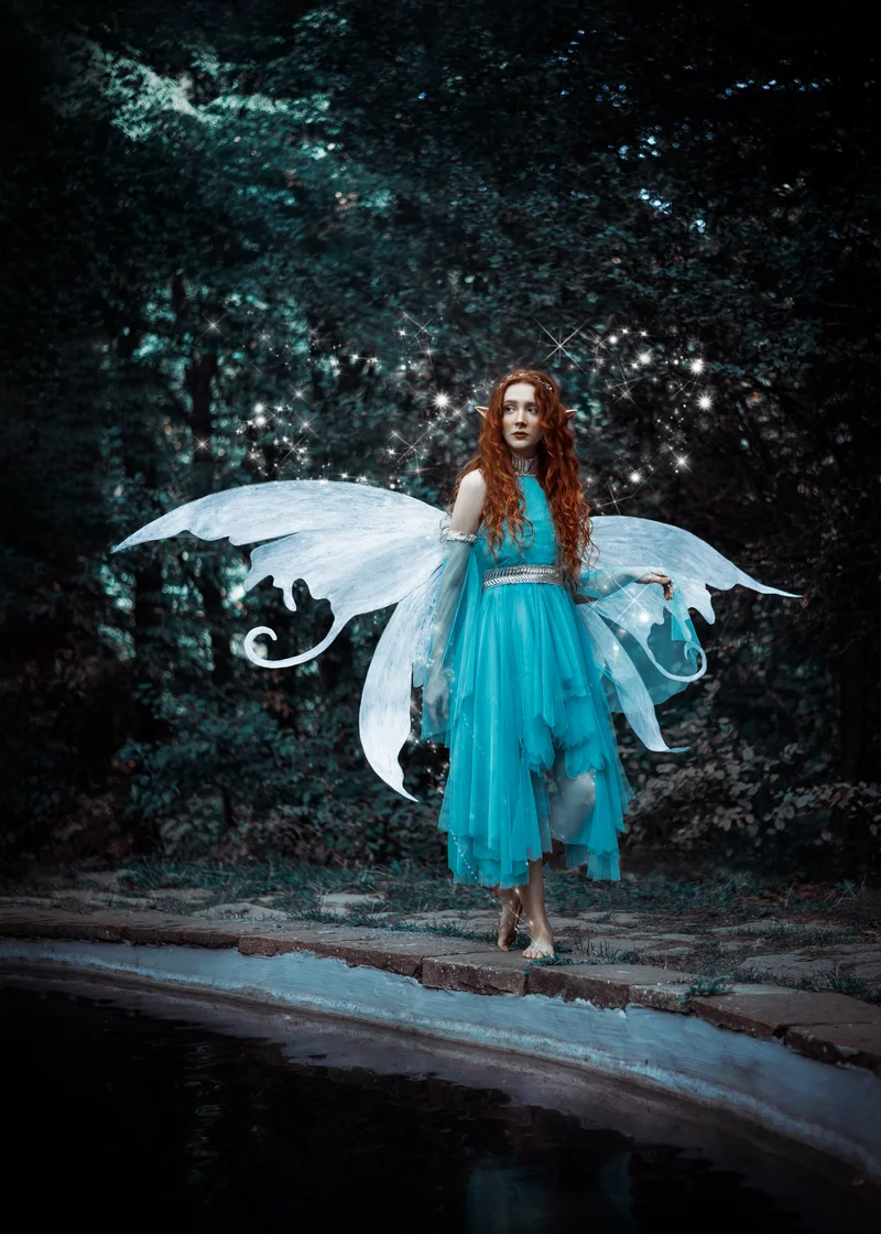 Red-haired woman with white fairy wings and a turquoise dress by a pond in the forest