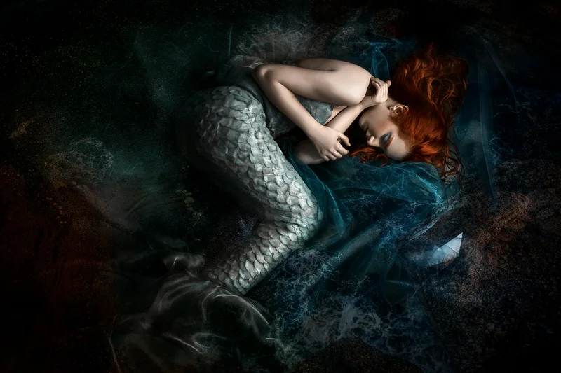 Red-haired woman as a mermaid with a silver tail, curled up against a dark background