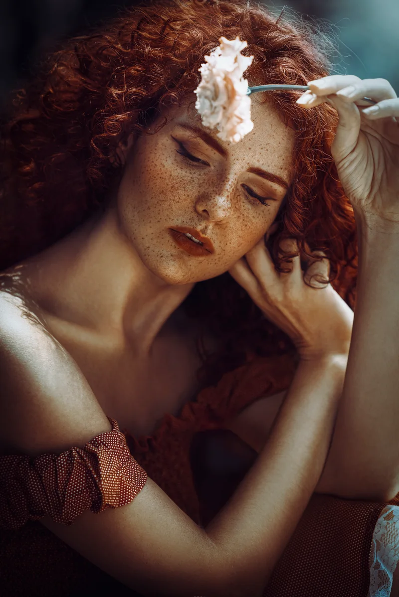 Freckled woman with curly red hair holding a white carnation above her brow